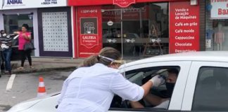 profissional de saúde com trajes de proteção se inclina para medir temperatura na testa de motorista dentro do carro, parado na avenida em frente a comércios