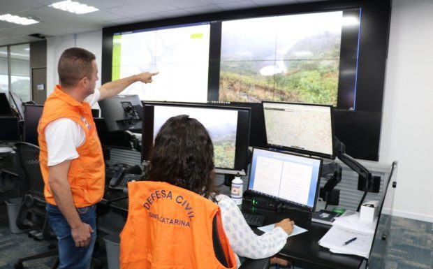 homem e mulher e mulher com coletes da defesa civil olhando para diversos monitores, em um deles há um rio seco