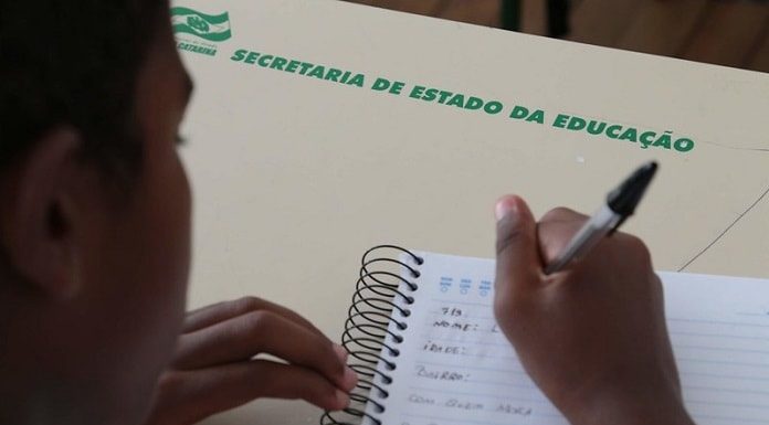 aluno escrevendo com caneta em caderno sobre carteira onde está escrito "secretaria estadual de educação"