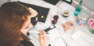 moça vista de cima com caneta na boca em frente à mesa com café e objetos de estudo