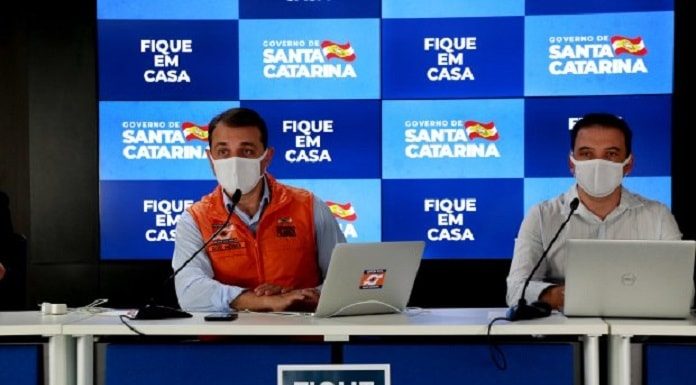 Carlos Moisés e Helton Zeferino em coletiva de imprensa online usando máscaras; painel de logos do governo ao fundo e mensagem "fique em casa"