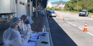 dois profissionais de saúde sentados em mesinha na plataforma do terminal à espera de carros; carros ao fundo; cones, viatura da guarda ao lado