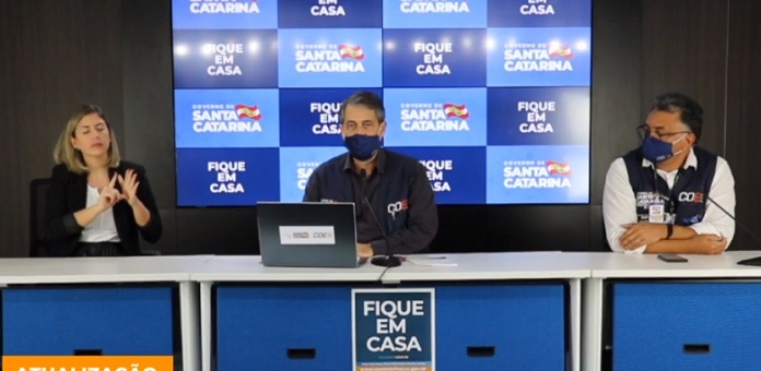 imagem da coletiva onde está, em uma bancada, o secretário ao centro, outro homem ao lado, e intérprete de libras do outro; painel de logos do governo ao fundo