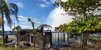 rancho destruído pelo incêndio, com fumaça saindo e algumas árvores em volta; bombeiro ao lado