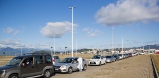 carros em fila no estacionamento externo do centro multiuso de são josé; profissional da saúde com trajes de proteção em pé ao lado