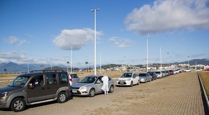 carros em fila no estacionamento externo do centro multiuso de são josé; profissional da saúde com trajes de proteção em pé ao lado