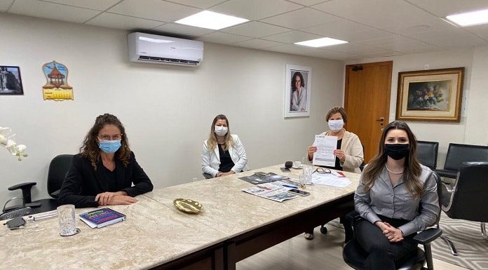 as quatro mulheres usando mascaras sentadas em volta de uma mesa olham para a câmera; adeliana mostra um papel