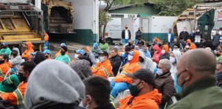 reunião de trabalhadores da comcap com uniformes, roupas de frio e máscaras, a maioria sentada, em pátio da empresa