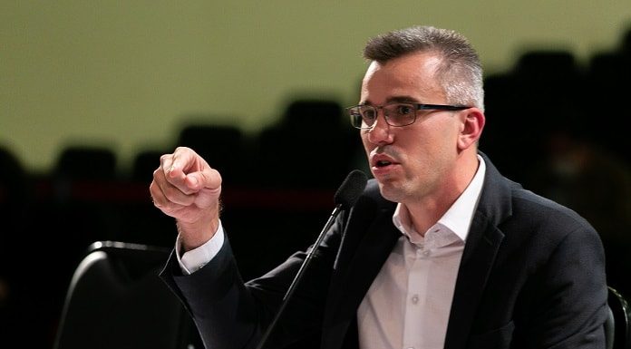 douglas borba fala ao microfone gesticulando; ele usa óculos e blazer; está em um auditório