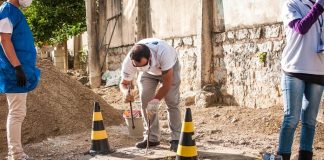 homem se abaixa para fazer inspeção em tampa de esgoto com cones em volta no meio de uma área em construção