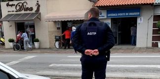 guarda de trânsito visto de costas com as mãos cruzadas para trás, olhando para rua, com carro estacionado ao lado