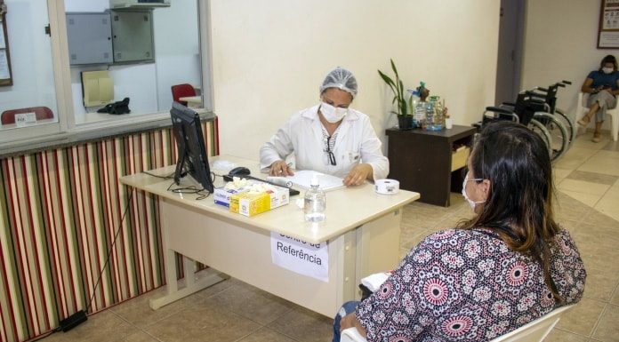 profissional de saúde com trajes de proteção sentada em mesa fazendo anotações e mulher em cadeira ao lado