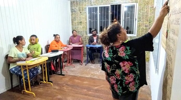 professora vista de costas dando aula apontando para o quadro em pequena sala com outras mulheres; há um garoto jovem com uma delas