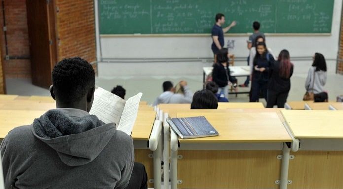 homem visto de costas em bancada de sala de aula com pessoas na frente da sala de aula, em pé em frente ao quadro negro