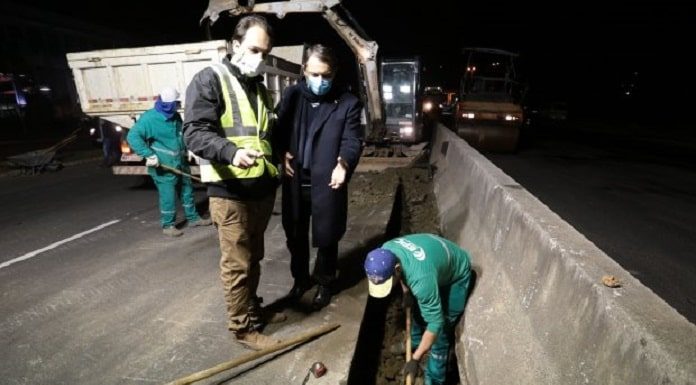 Dois homens de pé olhando um homem vestindo macacão verde, com boné azul, trabalhando com uma pá na mão