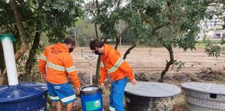 dois funcionários da comcap uniformizados carregam balde aparentemente pesado em pátio onde há caixas d'água espalhadas