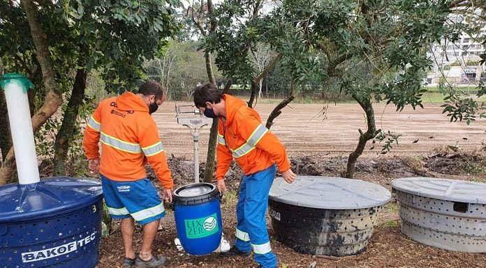 dois funcionários da comcap uniformizados carregam balde aparentemente pesado em pátio onde há caixas d'água espalhadas