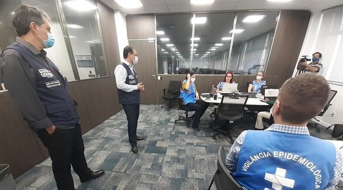 motta e moisés em pé conversando com servidoras com coletes do governo estadual em diferentes mesas em uma sala acarpetada; todos usam máscaras