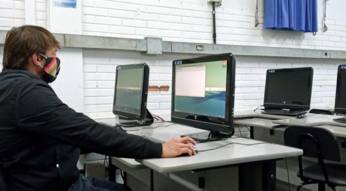 homem usando máscara sentado em frente a um computador em sala com mais computadores