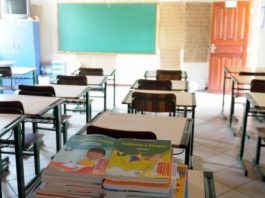 livros didáticos sobre uma carteira em sala de aula com outras mesas e carteiras organizadas; porta aberta ao lado do quadro