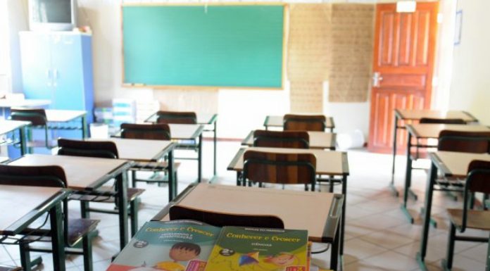 livros didáticos sobre uma carteira em sala de aula com outras mesas e carteiras organizadas; porta aberta ao lado do quadro