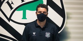 Homem vestindo camisa preta, usando máscara preta de pé em frente ao simbolo do Figueirense