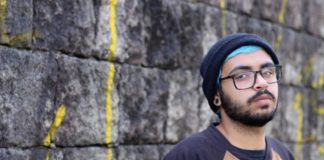 rapper warllock posa para a foto em frente a um muro; ele usa touca, óculos e tem barba