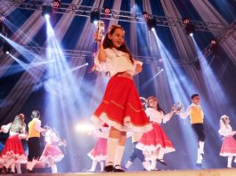 menina no palco em destaque no meio da foto dançando feliz com outras em volta com vestidos iguais; outros rapazes dançando junto e feixes de luzes azul