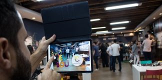 homem bate foto com tablet do ambiente do cocreation lab, onde há mesas e pessoas em pé no fundo