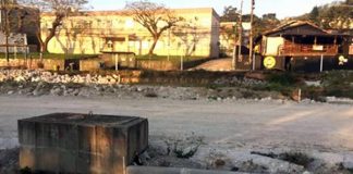 trecho pavimentado com britas e casas ao fundo. no meio da pavimentação há meio fio e caixa de concreto