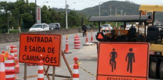 placas de sinalização de obra às margens da rodovia, indiciando entrada e saída de caminhões e passagem de pedestres e cilclistas; rodovias e carros ao fundo