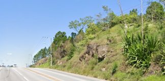 terreno íngreme com rochas e vegetação na margem da rodovia; carro ao fundo em cima do morro