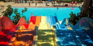 tobogãs coloridos em queda para uma piscina ao fundo, com banhistas circulando; sombra de árvore por cima do tobogã