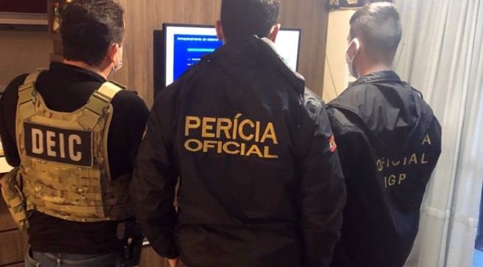 três agentes vistos de costas em pé em sala de apartamento olhando par uma televisão; as inscrições nos casacos de cada um são Dei, Perícia Oficial e Oficial IGP