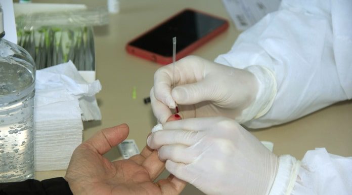 close em mãos de profissional de saúde aplicando teste na ponta do dedo de uma pessoa sobre uma mesa; profissional de saúde usa luvas e mangas compridas; não é possível saber seu gênero; materiais em cima da mesa