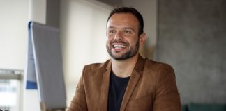 pedrão sorridente olha um pouco para o lado; ele está sentado em uma mesa de escritório, usando um blazer marrom