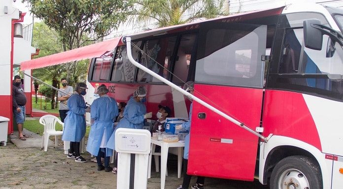 ônibus com toldo na lateral onde estão trabalhadoras usando trajes de proteção e manipulando os equipamentos de testes; homens de máscara ao fundo