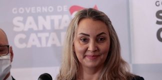 daniela reinehr em frente a um microfone com painel de logos ao fundo