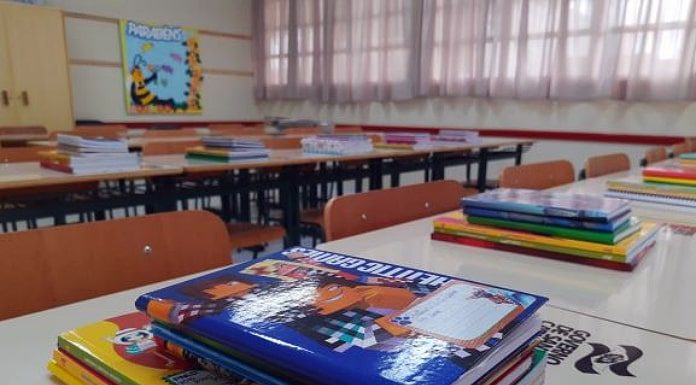 livros e cadernos organizados em pilha sobre carteira em sala de aula com mais mesas e carteiras organizadas; persianas fechadas