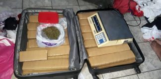 mala aberta do chão com grandes tabletes empacotados e uma sacola por cima com maconha; no outro lado uma balança