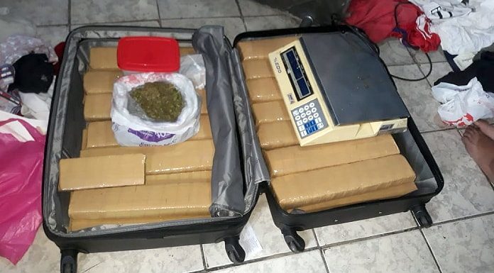 mala aberta do chão com grandes tabletes empacotados e uma sacola por cima com maconha; no outro lado uma balança