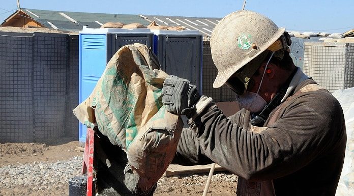 trabalhador da construção civil usando máscara, capacete e óculos de proteção faz preparo de cimento em cima de bancada em área de construção