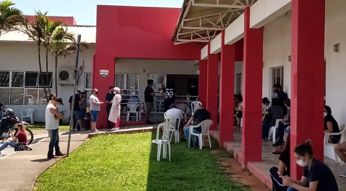 gramado com cadeiras em frente ao cati onde há pessoas em volta usando máscaras esperando em filas e sentadas em volta