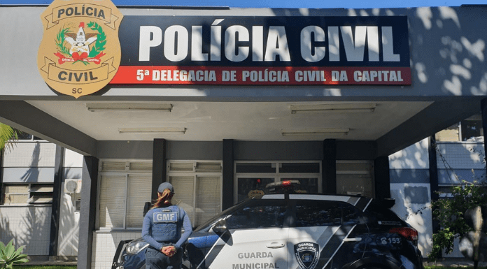 Fachada da Delegacia da Polícia Civil, com um carro da Guarda Municipal e uma policial com colete da GMF à frente