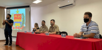 Aula inaugural da Disciplina de Libras, um intérprete em pé em frente à uma grande mesa com toalha vermelha, onde estão sentados a vereadora Cristina de Souza, os coronéis Dionei Tonet e Fábio José Martins, e o professor Deonísio Schmitt da UFSC. 