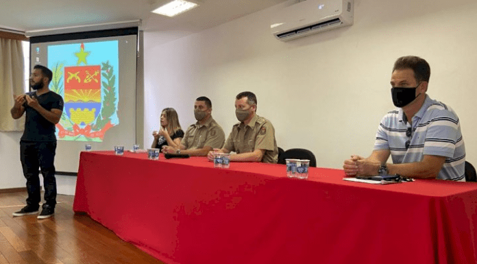 Aula inaugural da Disciplina de Libras, um intérprete em pé em frente à uma grande mesa com toalha vermelha, onde estão sentados a vereadora Cristina de Souza, os coronéis Dionei Tonet e Fábio José Martins, e o professor Deonísio Schmitt da UFSC. 