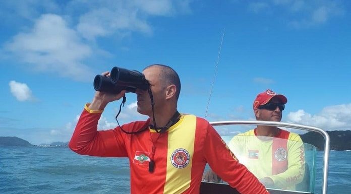 em uma lancha, bombeiro usa binóculos olhando para o mar, outro ao fundo pilota o barco