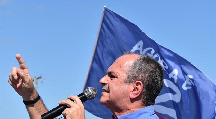 moacir visto de lado discursando ao microfone com dedo em riste; bandeira com seu número ao fundo