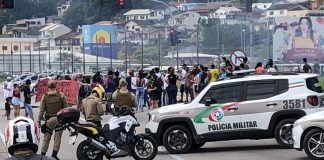 carro jeep da polícia militar atressado na pista com moto lado; motoqueiros em primeiro plano no trânsito. ao fundo no centro da foto grupo protestando e alguns policiais em volta; morro com casas ao fundo