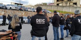 Mecanismo verde: diversos policiais civis homens e mulheres com jaquetas da corporação reunidos em pé em estacionamento; maioria de costas para foto